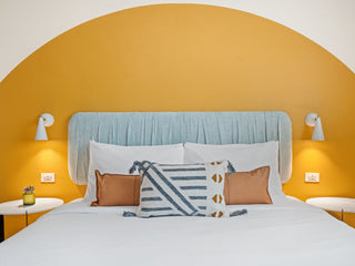 Hotel room with bed covered in white linens with gold half circle painted behind and sconce lights on either side.