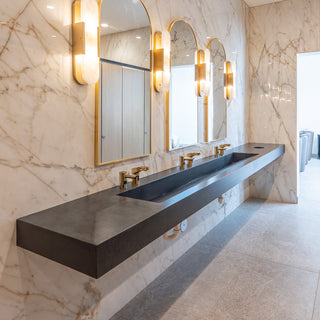 Black concrete triple station commercial sink in ramp style against a marble wall with gold framed mirrors and sconces.