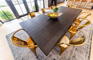 Dark wood look concrete dining table with a beveled edge, paired with metallic gold industrial-style chairs in a sunlit modern dining room—ideal for luxury residential interiors, hospitality spaces, and statement-making design projects.