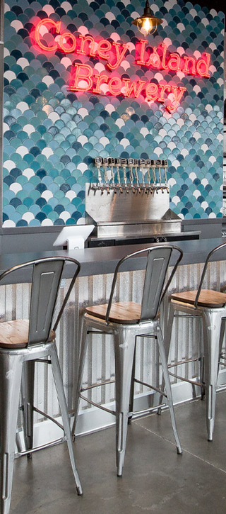 Bar with gray concrete countertops and blue fan shaped tile wall. Neon sign states Coney Island Brewery.